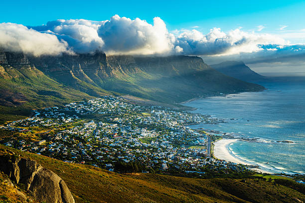 A sweeping coastal cityscape of Cape Town from Lions Head overlooking Camps Bay, with Table Mountain and the Twelve Apostles in the distance. Bright blue skies, sparkling sea, and urban rooftops create a vibrant, scenic panorama.