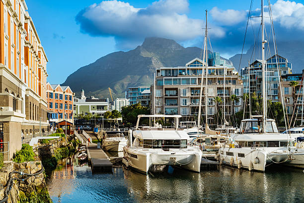 A vibrant Cape Town harbor scene featuring luxury yachts, calm water, and a backdrop of Table Mountain and Twelve Apostles. Modern apartment buildings line the marina under a blue sky.