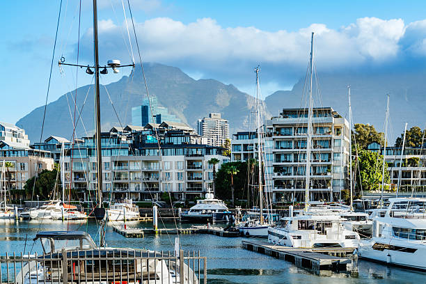 A sunny Cape Town harbor scene featuring sailboats and yachts moored in a marina, with modern waterfront apartments and the Twelve Apostles mountains in the distance.