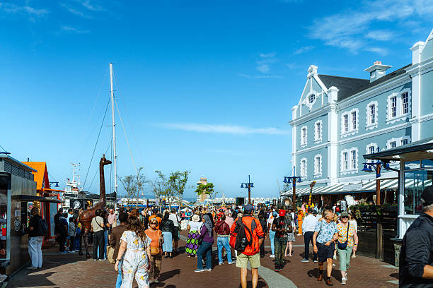Crowded Cape Town waterfront street lined with historic buildings and outdoor cafés, tourists and locals mingle under a bright blue sky near the harbor, with mountains in the distance.