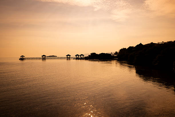 Silhouetted wooden huts along a jetty at sunset on Seraya Island in Komodo National Park, Indonesia, with warm golden light reflecting on calm water and a peaceful tropical atmosphere.