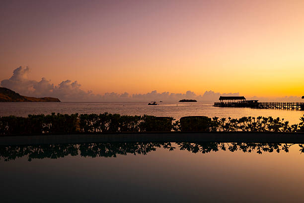 Sunset over a wooden jetty on Seraya Island in Komodo National Park, Indonesia, with warm evening light reflecting on calm water and simple huts silhouetted against the horizon.