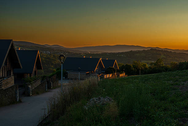 Lanckorona, Poland – August 19, 2025: Warm sunset light silhouettes distant mountain ridges behind hillside homes and grassy slopes. Atmospheric evening scene for travel, rural lifestyle, and peaceful neighborhood concepts with dramatic sky gradient.