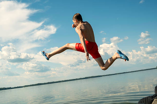Energetic young man performs a joyful leap into a calm lake, showcasing exuberance and freedom. The vibrant blue sky and fluffy clouds set a perfect summer backdrop for this action shot.