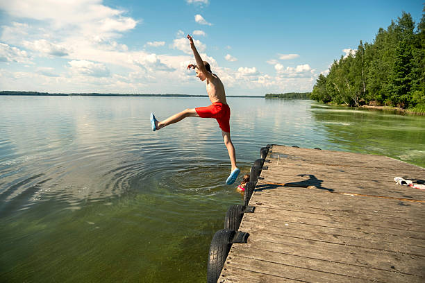 Athlete Young men  diving into water from wooden dock on a lake. Eco tourism  Summer refreshment by the water of a lake in nature. Relaxing and enjoying the lakeside, doing water sports. Beautiful nature sky and lake landscape in background.