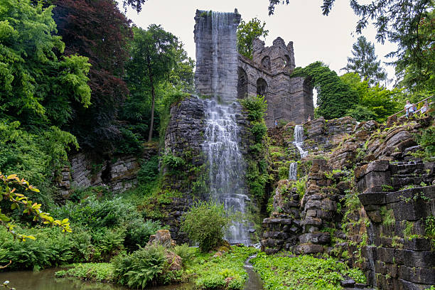 Waterfall Closeup Over Mossy Stone Arch With Vibrant Yellow Flowers And Lush Greenery Powerful Spray Drifts Across Camera, Textured Stone And Fern Fronds Frame Foreground. Kassel, Germany.