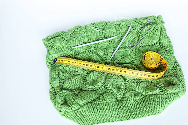 Green hand-knitted sweater with a distinctive cable pattern; knitting needles, a stitch holder, and a yellow measuring tape on top highlight craftsmanship, creativity, traditional knitting, hobbies, and handmade crafts.