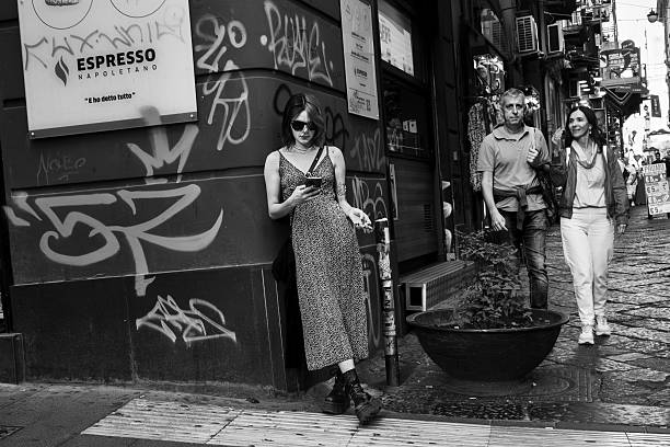 Black and white editorial photograph of a young woman leaning against the wall on Via Toledo in Naples while using her phone. The image captures the city's daily life, with buildings, passersby, and the crowd in the background. A realistic, spontaneous, and dynamic urban shot.