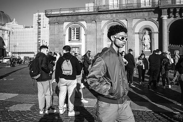 Young man walking in Piazza Pebliscito, Naples. The image captures the city's daily life, with buildings, passersby, and the crowd in the background. A realistic, candid, and dynamic urban shot.