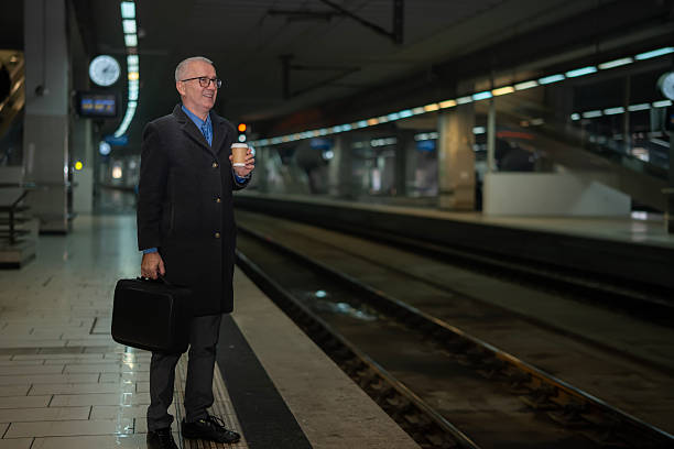 Mature adult businessman holding a disposable coffee cup and briefcase, smiling while waiting on a metropolitan train station platform for a departure