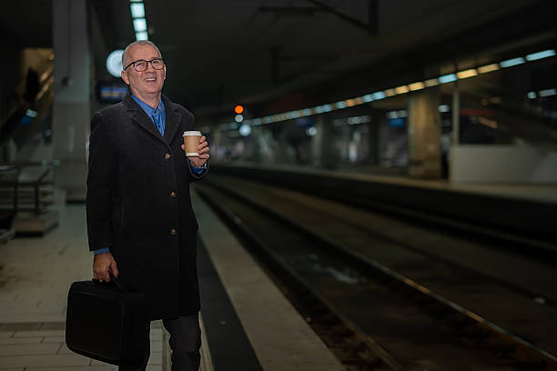 Mature adult man standing on an underground train station platform at night, holding a disposable coffee cup and a briefcase while waiting for a train