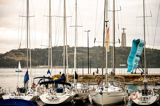 Lisbon Portugal December 06 2025. Multiple sailboats are docked at a riverside marina along the Tagus River with Cristo Rei monument visible across the water.