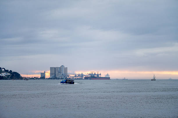Lisbon Portugal December 06 2025. A small passenger boat crosses the Tagus River with industrial port structures visible in the distance at dusk.