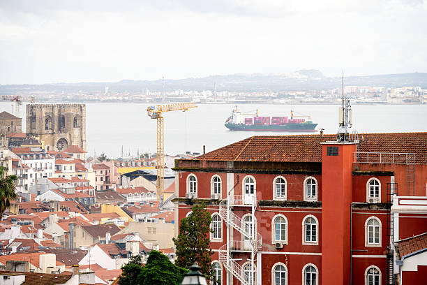 Lisbon Portugal December 06 2025. A red building with an exterior spiral staircase overlooks Lisbon rooftops with a cathedral and a cargo ship on the Tagus River.