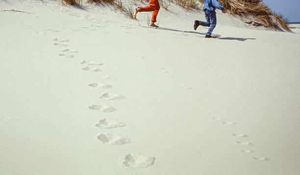 Two people running one behind the other across pale sand dunes, leaving a trail of footprints in the sand. A minimalistic and dynamic image symbolizing freedom, movement and carefree outdoor activity. Suitable for creative and conceptual use in lifestyle, nature, travel and outdoor themes.