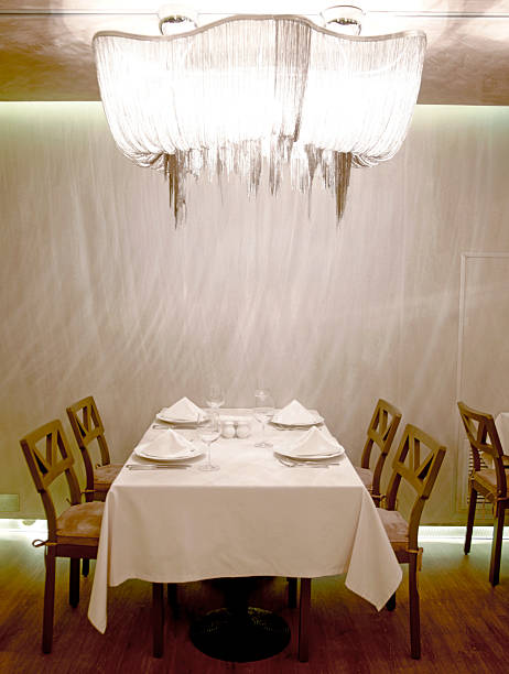 Vertical full frame image of a sophisticated restaurant interior featuring a neatly arranged table with white tablecloth, classic chairs, and a sparkling crystal chandelier hanging overhead. The warm ambient lighting and soft neutral wall textures create an intimate modern interior scene with copy space that conveys luxury dining and upscale hospitality. This stylish space is ideal for showcasing high end restaurants, romantic dinners, and exclusive event venues in editorial and commercial visuals.