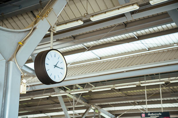 Analog clock hanging inside modern train station with industrial roof structure. Concept of time management, punctual schedule, public transportation, waiting journey and urban commuting lifestyle.