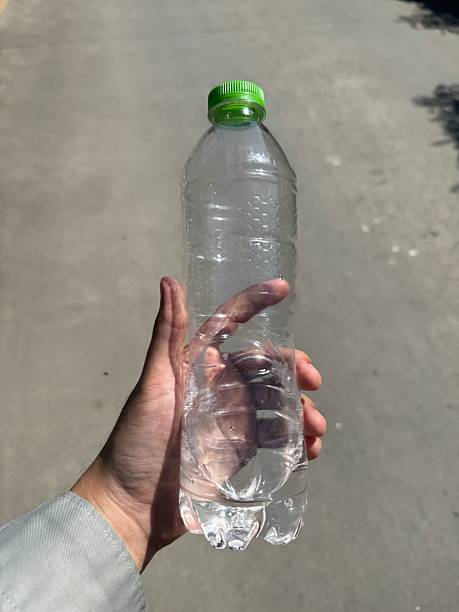 istock person's hand holding a clear, empty plastic water bottle with a bright green cap outdoors. The bottle features a hexagonal textured pattern. 2253338164