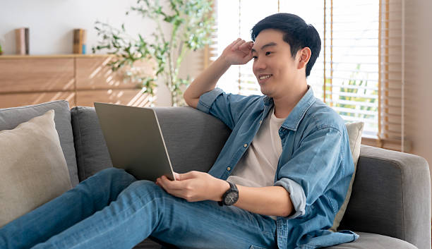 istock Young Asian man relaxing on comfortable couch, using laptop at home, happy boy chatting with friends in social network, spending weekend, watching movie, shopping online 2253338304