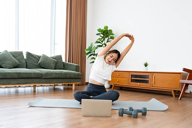 istock Young Asian woman stretching yoga workout on exercise mat while online training class with computer laptop in living room 2253338327