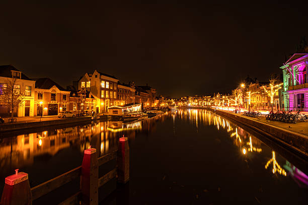 Atmospheric night cityscape of Haarlem, the Netherlands, seen from the Gravenstenen Bridge over the Spaarne River. Historic buildings are decorated with festive Christmas lights, while long exposure captures colorful light trails from passing cars and warm reflections in the calm water, creating a vibrant yet serene holiday atmosphere.