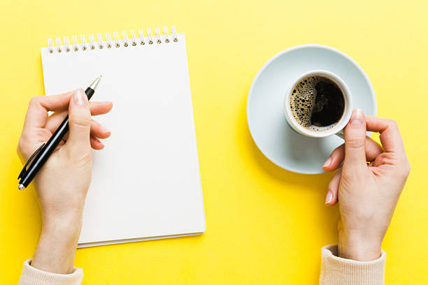 Woman hand with pencil writing on notebook and hold coffee cup. Woman working on office table with coffee.