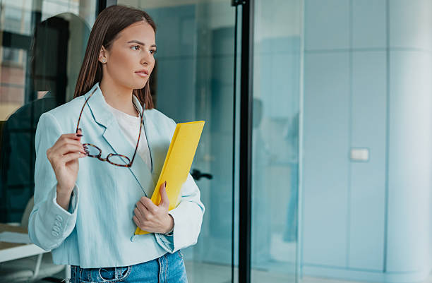 istock Office worker holding files and glasses while standing near a glass wall in a modern office setting during the day 2253352401