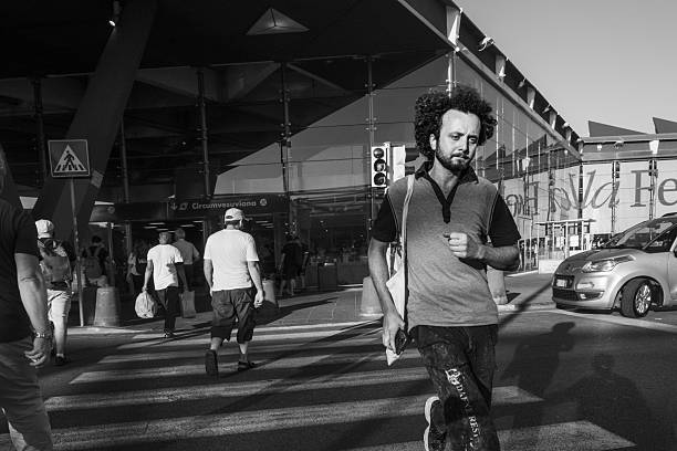 Naples, Italy – May,  2022.: A man walks near the exit of Naples Central Station in a busy urban area. The black-and-white photograph shows pedestrians crossing the street, vehicles in traffic, and the station building in the background, capturing the daily hustle and bustle of life in the city's transportation district.
