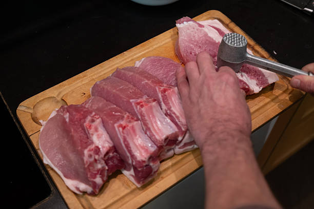 A man tenderizes fresh raw pork chops with a metal mallet on a wooden cutting board. The process of cooking a homemade meal in the kitchen.