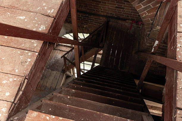 A dusty, weathered wooden staircase winds downward through a brick attic, creating a moody, vintage scene. Narrow steps, rough beams, and muted light evoke mystery, history, and quiet exploration within an old building