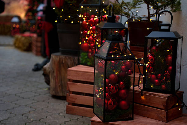 festive outdoor display featuring black lanterns filled with red christmas ornaments and string lights, situated on wooden crates, creating a warm and inviting holiday atmosphere, greenery