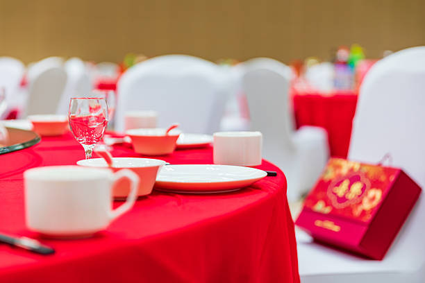 A close-up of clear tall candle holders and lush floral arrangements in yellow and white that run along the table. In the blurred background are round banquet tables with white chair covers and gift bags, warm lighting producing a soft glow. The shallow depth of field emphasizes the texture and colors of the centerpiece while conveying an elegant, festive mood.