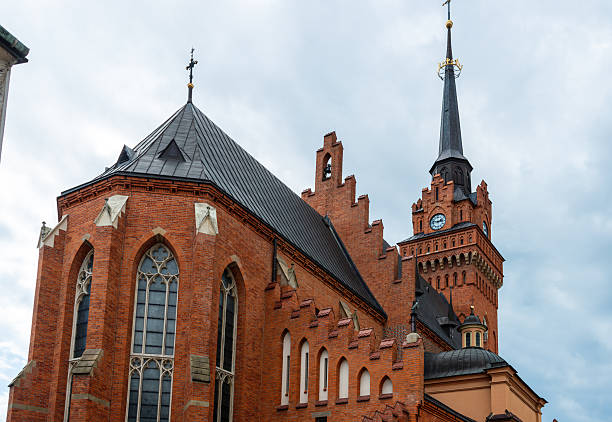 Tarnow, Poland – July 12, 2025: Red brick Gothic church facade with tall clock tower and pointed spire rises against a cloudy summer sky. Architectural details emphasize the historic religious character of the city.