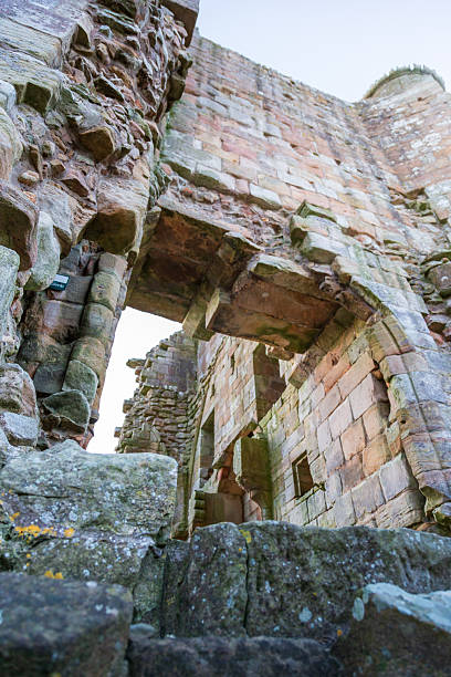 Edlingham Castle, located in Northumberland, is depicted with its ancient stone walls and arches partially intact. The structure is constructed from rough-hewn stones, showcasing a blend of reddish and grey hues. The castle, seen in the soft light of dawn, reveals the weathering from centuries. Moss and lichen grow on the stones, adding texture and character. The lack of roof and partial walls give a glimpse of its historical past, set against a clear sky, typical of a winter morning.