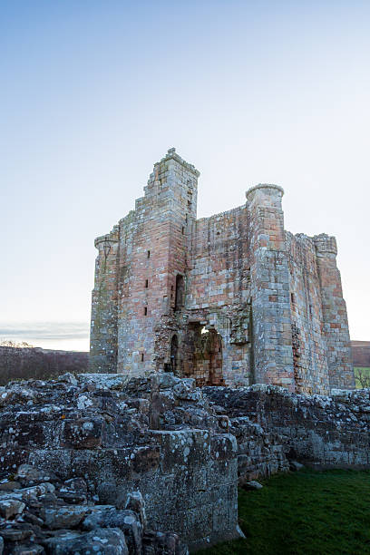 Edlingham Castle in Northumberland is depicted at dawn during winter. The ruins display a robust stone structure with visible archways and battlements, showcasing medieval architecture. The castle is built with weathered stones, creating a textured facade. The surrounding environment features a grassy area and rugged stone wall remnants. The sky is clear, offering a soft backdrop that enhances the ancient feel of the site. The overall ambiance is serene, with historical significance amidst the natural landscape.