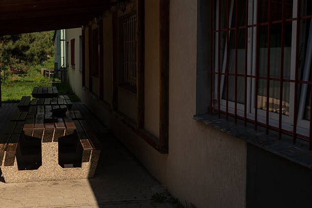Shaded picnic benches under covered walkway with barred windows and concrete table. Wooden slat seating faces grassy yard, low light and deep shadows