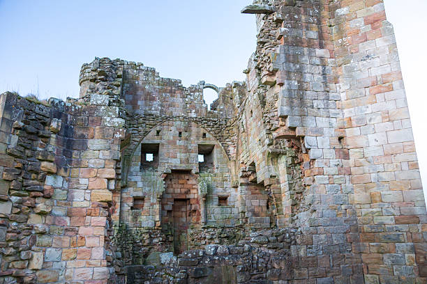 Ancient stone walls of Edlingham Castle in Northumberland, UK, seen at dawn during winter. The irregular, weathered stones form the standing remains of the medieval fortress. Large archways and small windows punctuate the structure, reflecting the castle's historical architecture. The sky is clear, casting a soft light on the castle, highlighting its textured and varied stonework. The structure is surrounded by natural growth, typical of the region’s historical sites.