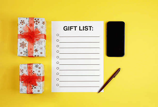 Flat lay a gift list, smartphone, and pen on a yellow background. Festive holiday planning, Christmas online shopping, and New Year preparation.