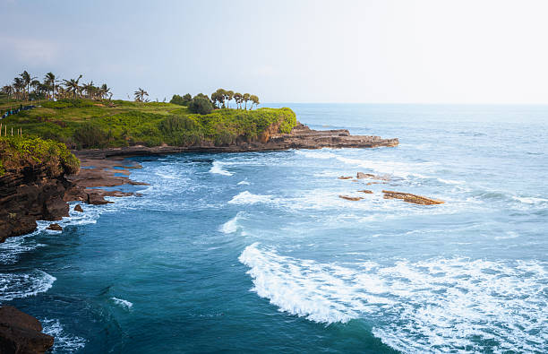 Tanah Lot coastline dramatic ocean cliffs and tropical shore near Bali temple area create calm travel mood with blue water, volcanic rocks, Bali coastal nature and open horizon under soft daylight.