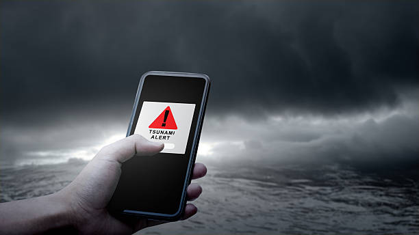 istock Hand holds a smartphone displaying a tsunami alert message against a dramatic backdrop of dark, ominous clouds and turbulent ocean waters. Technology in disaster warning and emergency preparedness 2253397687