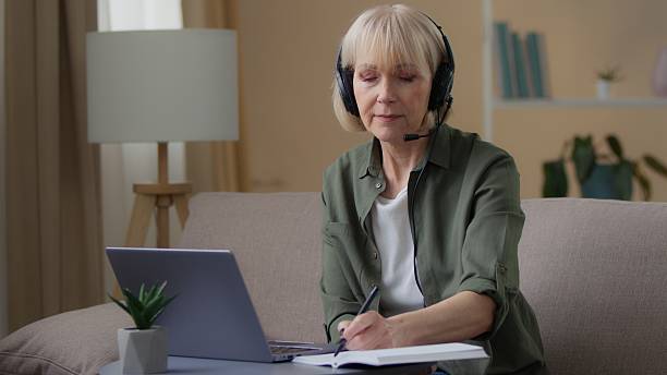 istock Senior old Caucasian woman in headphones writing notes watching webinar e-learning at home retired female write information looking in laptop computer on couch listen online training video conference 2253398118