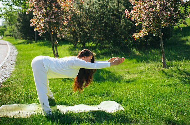 istock A woman in a white outfit performs a forward bend yoga pose on grass, framed by cherry blossoms and bathed in warm sunlight. 2253398704