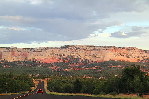 A lone vehicle travels down a long, paved highway toward a backdrop of layered red and white rock mountains under a dramatic cloudy sky.