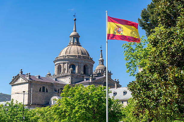 A Spanish flag flies before the grand dome and granite walls of El Escorial monastery in San Lorenzo, Spain, surrounded by green trees under a bright blue sky