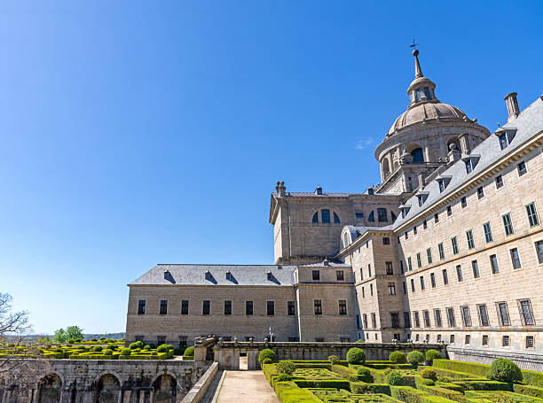 A side view of El Escorial in Spain, showing the geometric boxwood gardens, the long granite facade with slate roofs, and the central basilica dome under a blue sky