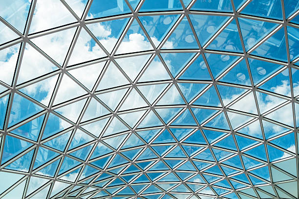 Glass ceiling with geometric pattern reflecting a vibrant blue sky and scattered clouds. The architectural design creates a modern, airy, and open ambiance in the space.