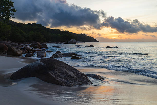 A peaceful coastal scene at sunset features smooth sand, large rocks along the shore, and gentle waves. Warm sky hues reflect on wet sand of Anse Lazio beach, Praslin island, Seychelles