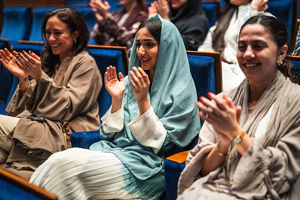 Young Middle Eastern female university students in Riyadh expressing enthusiasm by clapping and smiling during an inspiring panel discussion and recognition event in an auditorium.