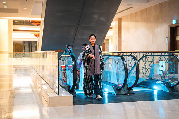 Middle Eastern female students walking through a modern campus building at Riyadh University, engaging in conversation while using an escalator, with bright interior lighting enhancing the academic environment.