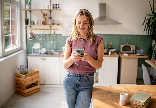 istock Young woman enjoys texting while standing in a bright kitchen during a sunny day in her apartment 2253409491